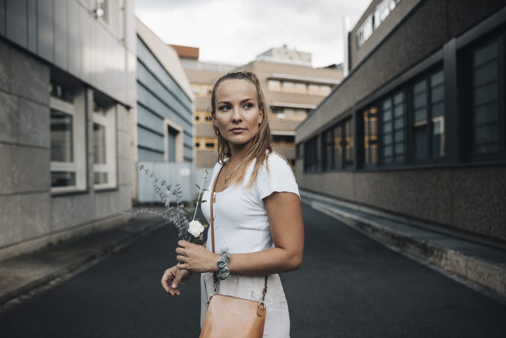 This is me, German Photographer based in Würzburg and Nuremberg, Bavaria. I am standing in the middle of a street wearing everyday clothes looking away from the camera, holding flowers in one hand. I am wearing a high ponytail with my blonde hair.