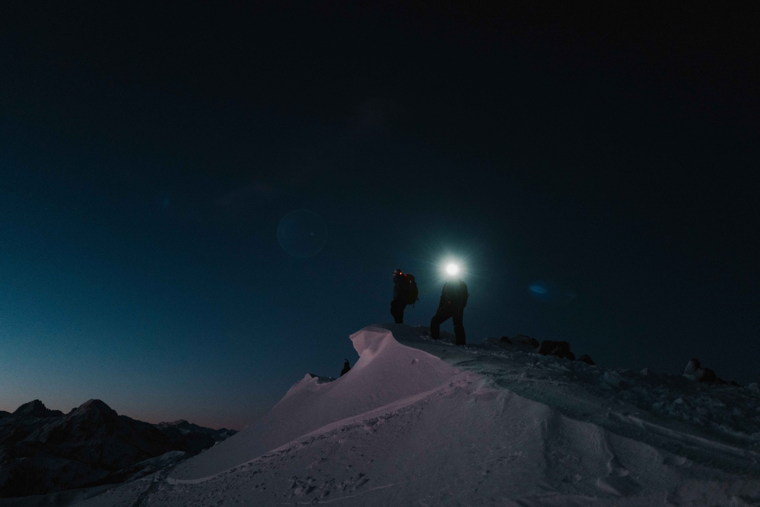 Two mountain climbers standing on top a mountain edge with strong headlamps.